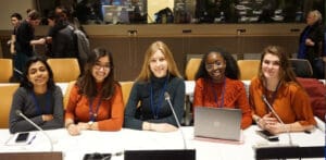 Five young women sitting behind a desk in a busy professional environment.
